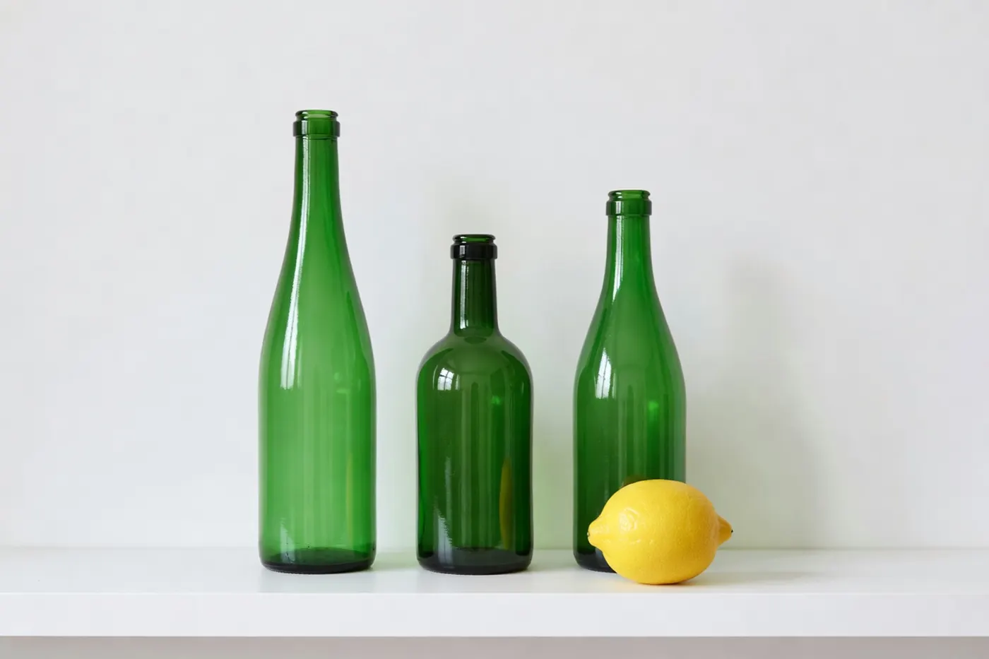 Grok Imagine Image: Exactly three green glass bottles standing on a white shelf. The tallest bottle on the left, the shortest in the middle, and a single yellow lemon on the shelf in front of the right-hand bottle. Plain white wall, soft even light.