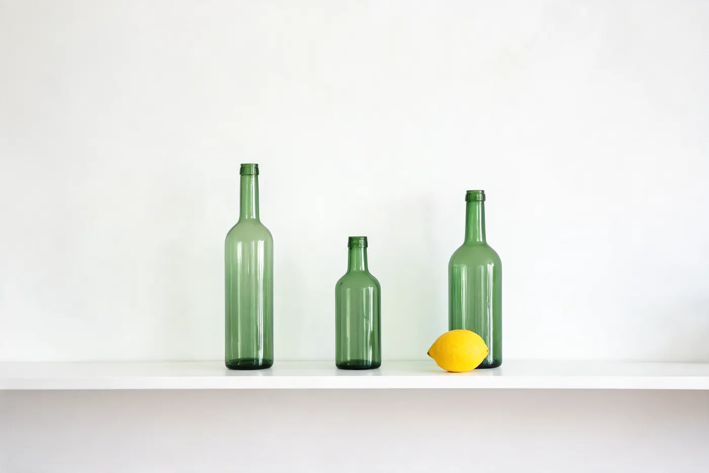 Seedream 5 Lite: Exactly three green glass bottles standing on a white shelf. The tallest bottle on the left, the shortest in the middle, and a single yellow lemon on the shelf in front of the right-hand bottle. Plain white wall, soft even light.